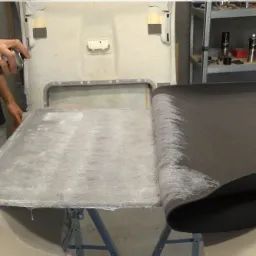 Przyklejanie czarnej tkaniny do podsufitki samochodowej za pomocą kleju w sprayu, widoczny fragment nieprzyklejonej tkaniny i osoba z puszką kleju.