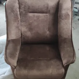 Brązowy fotel tapicerowany aksamitnym materiałem, widoczny z przodu, na podłodze leży biała tkanina, w tle warsztat z narzędziami.