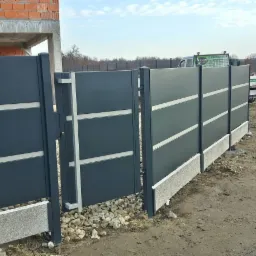 Nowoczesna, grafitowa brama wjazdowa z poziomymi, jasnymi akcentami, zamontowana na betonowej podmurówce, widok z zewnątrz na tle budowy i polnej drogi.