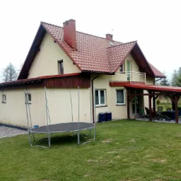 Dwupiętrowy dom z beżową elewacją i czerwoną dachówką, widoczny z ogrodu, z trampoliną na pierwszym planie i zadaszonym miejscem do siedzenia z tyłu domu.