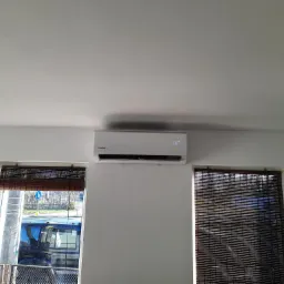 Biały klimatyzator Kaisai zamontowany na ścianie nad oknami z bambusowymi roletami, widoczny wyświetlacz z temperaturą 26 stopni.