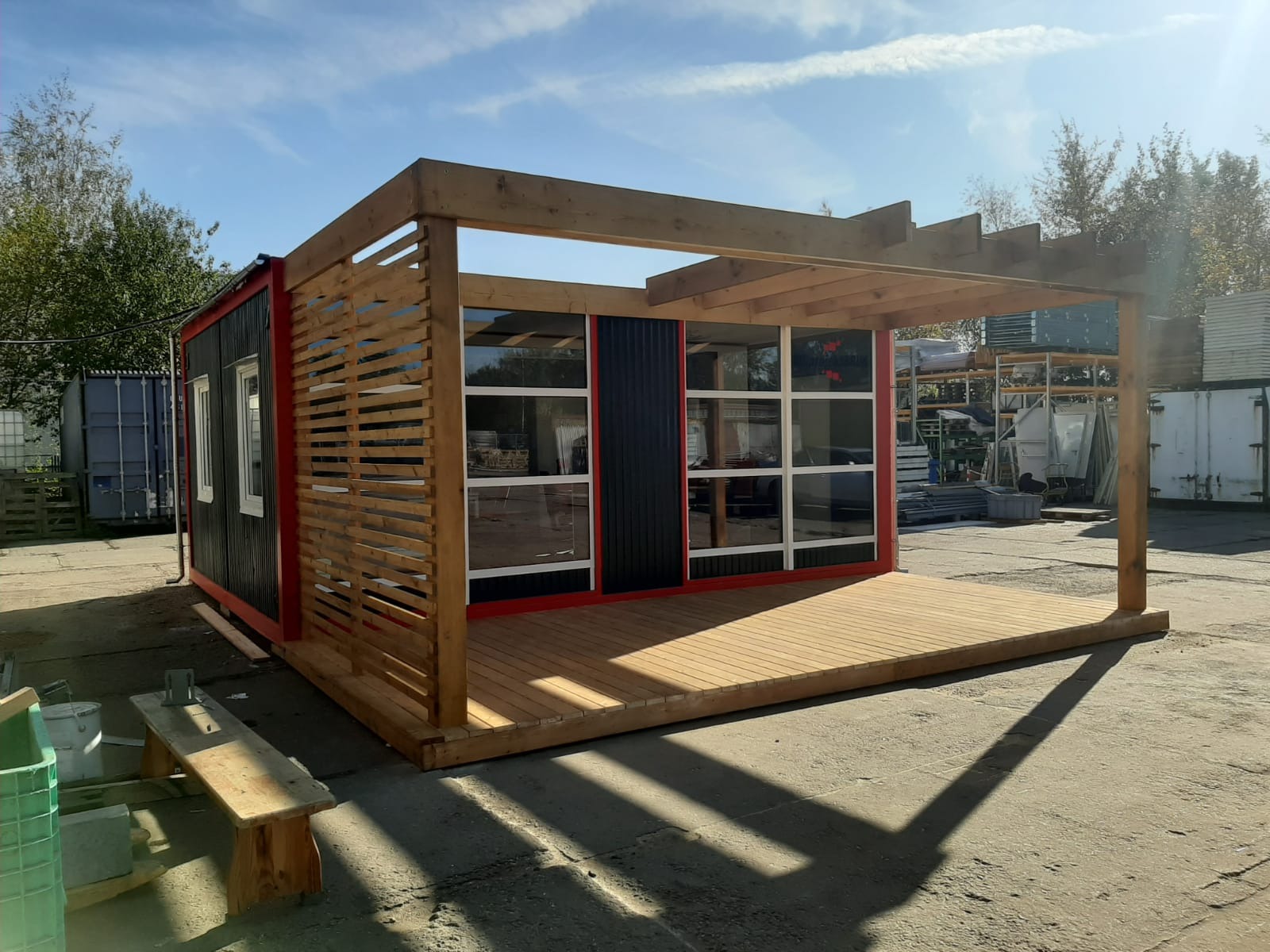 Nowoczesna altana ogrodowa z drewnianym tarasem i ażurową ścianą, zbudowana z kontenera, pomalowana na czarno z czerwonymi akcentami, w tle widoczne magazyny i błękitne niebo.