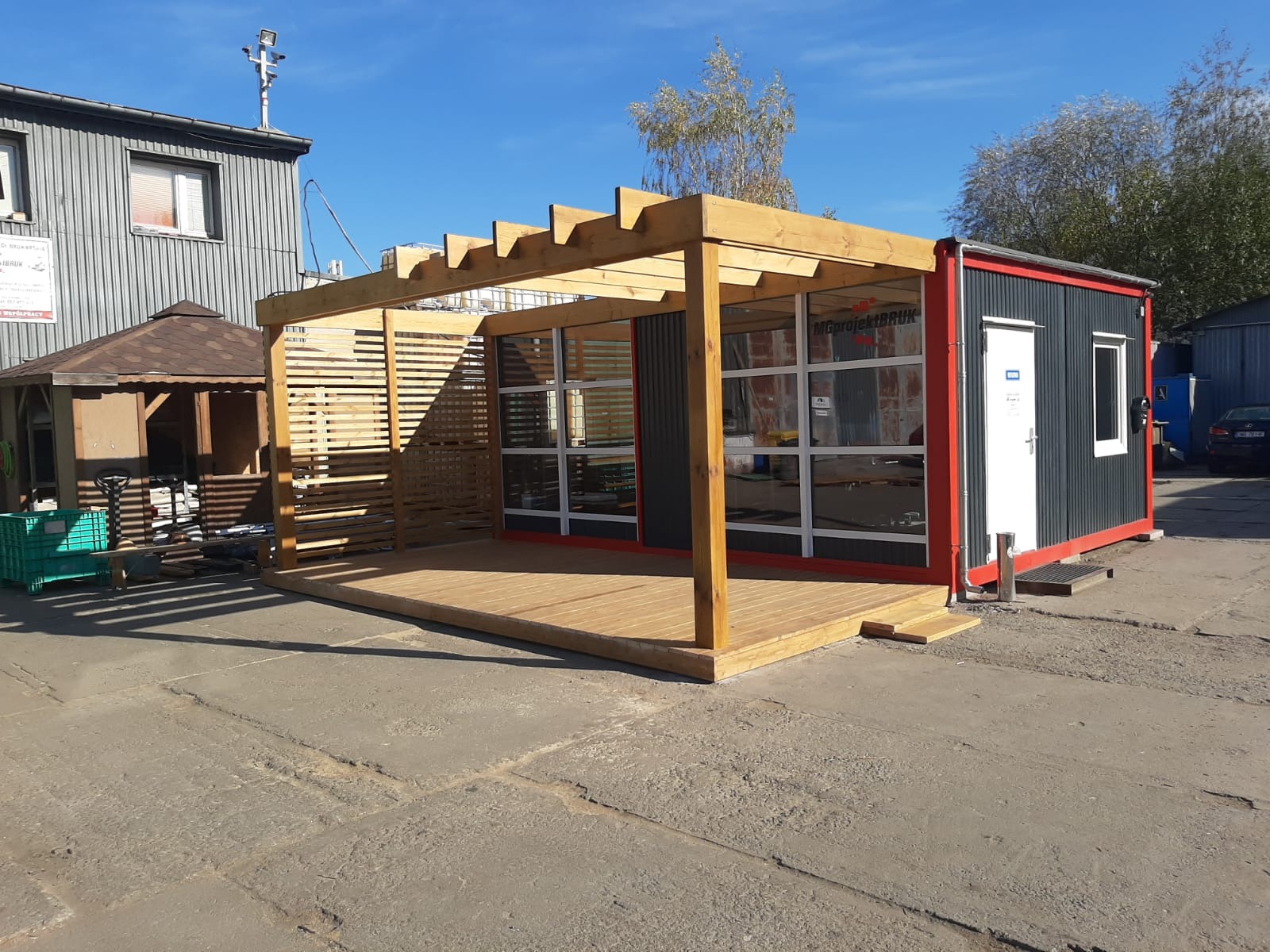 Nowoczesna konstrukcja z drewna i kontenera: taras z pergolą, zabudowa z przeszklonymi ścianami i wejściem, szara elewacja kontenera z czerwonymi akcentami, widok na plac.