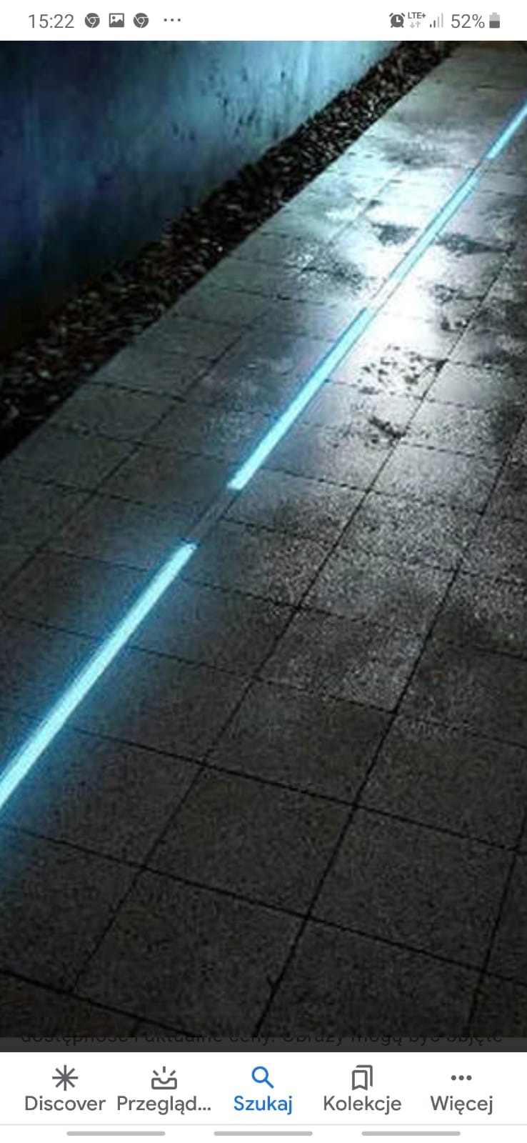 Szary chodnik z kostki brukowej z wbudowanym, podłużnym oświetleniem LED o chłodnej, niebieskiej barwie, oświetlający ścieżkę wzdłuż ciemnej ściany z dekoracyjnym żwirem.