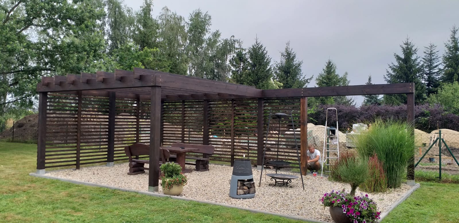 Drewniana pergola z poziomymi listwami, betonowa obwódka, żwirowe podłoże, drewniany stół z ławkami, palenisko z drewnem, drabina i mężczyzna w tle, rośliny ozdobne w donicach.