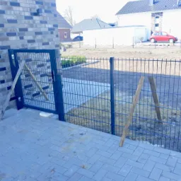 Montaż metalowej bramki w ogrodzeniu panelowym, podpartej drewnianymi stemplami, na tle nowego domu i przygotowanego terenu pod trawnik.