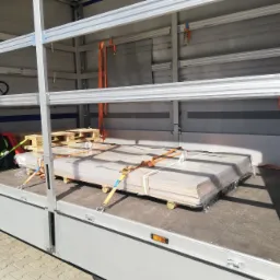 Ładunek płyt drewnianych zabezpieczony pasami transportowymi wewnątrz aluminiowej naczepy z widocznym wózkiem paletowym.