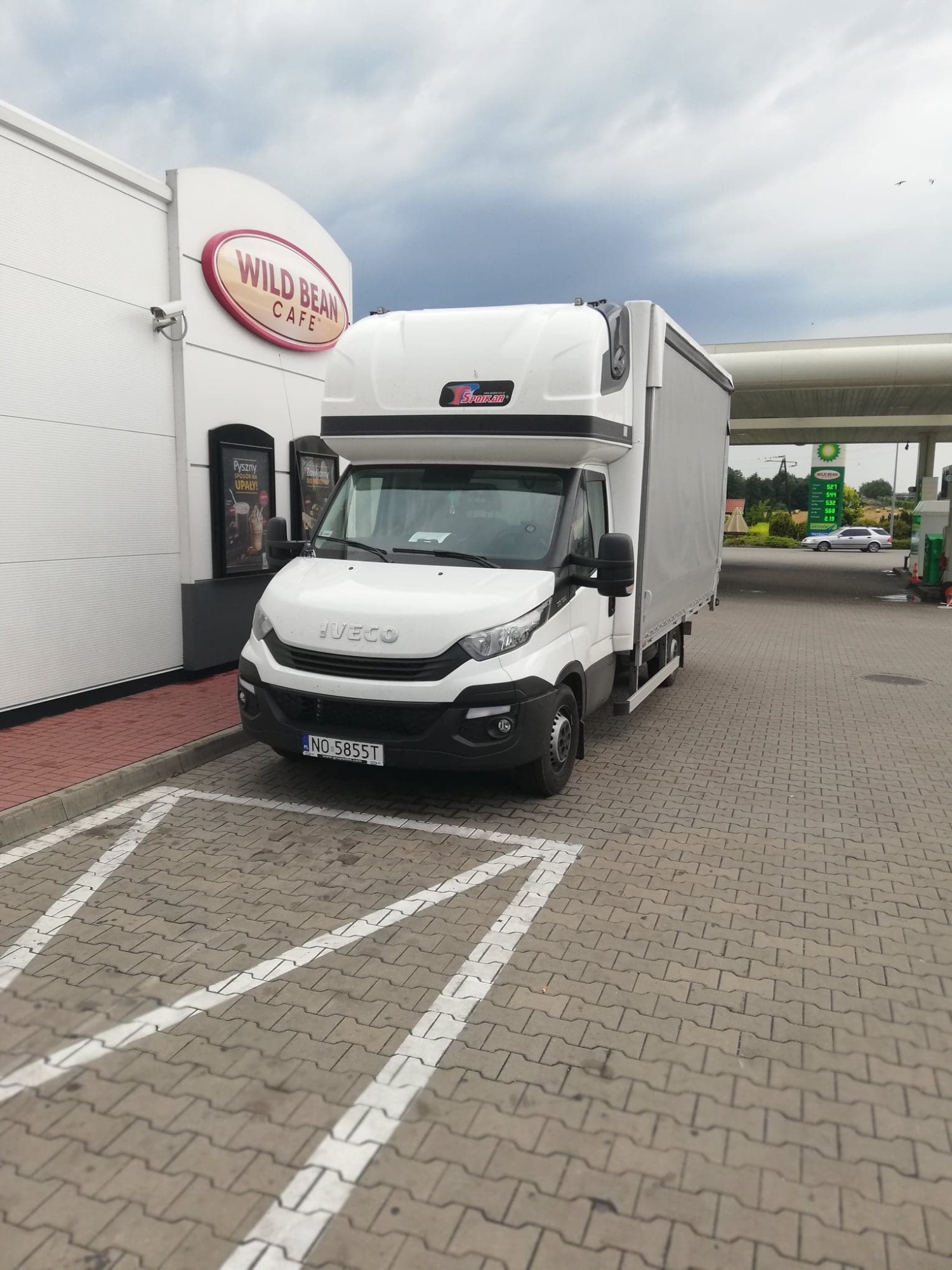 Biały samochód dostawczy Iveco z plandeką na parkingu stacji benzynowej, obok kawiarni Wild Bean Cafe. Widoczny numer rejestracyjny NO 5855T.