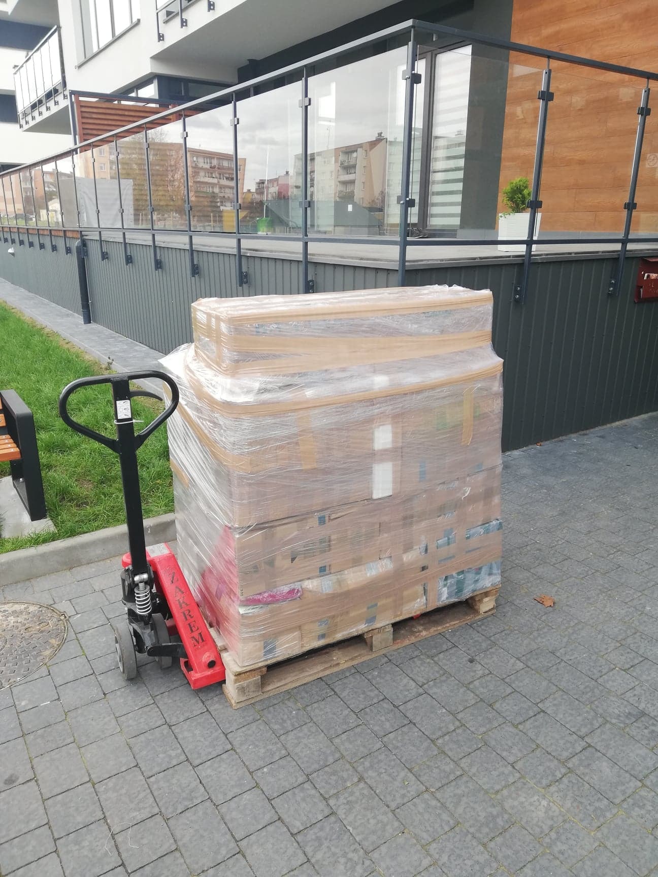 Paleta z kartonami owinięta folią stretch, transportowana wózkiem paletowym przed nowoczesnym budynkiem mieszkalnym z przeszklonymi balkonami.