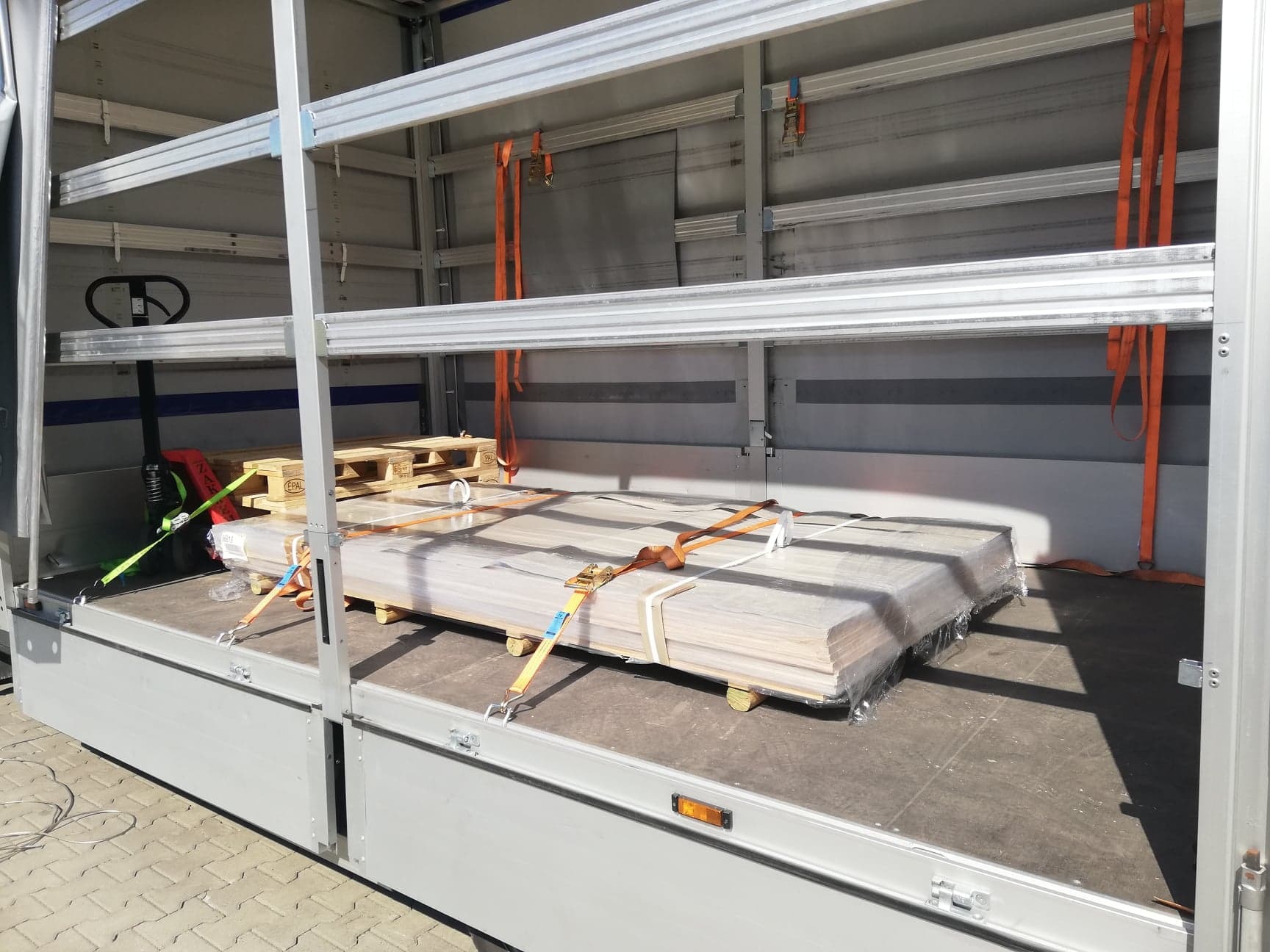 Ładunek płyt drewnianych zabezpieczony pasami transportowymi wewnątrz aluminiowej naczepy z widocznym wózkiem paletowym.
