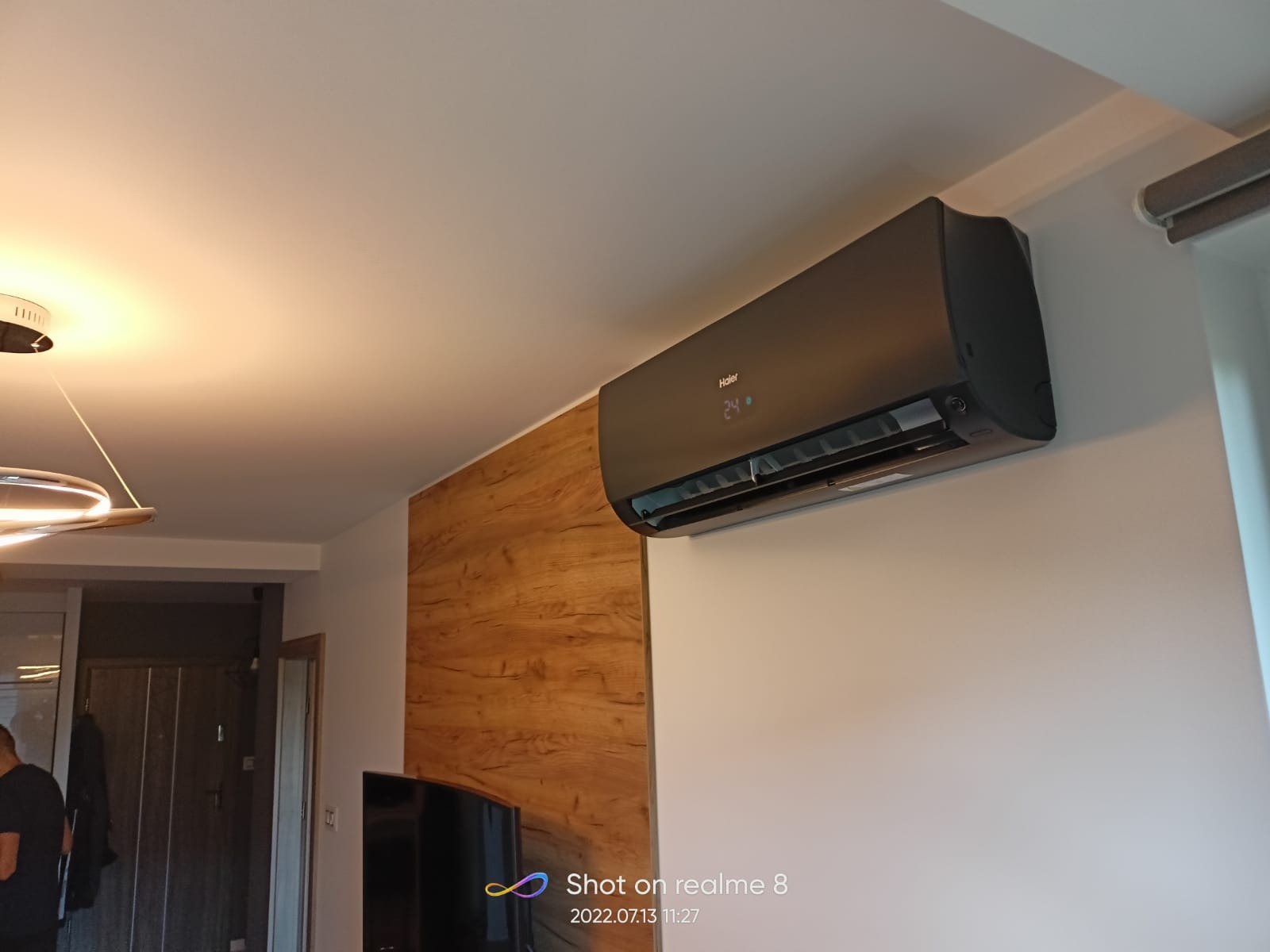 Czarna klimatyzacja Haier zamontowana na białej ścianie w nowoczesnym salonie z drewnianą okładziną. Na wyświetlaczu widoczna temperatura 24 stopnie.