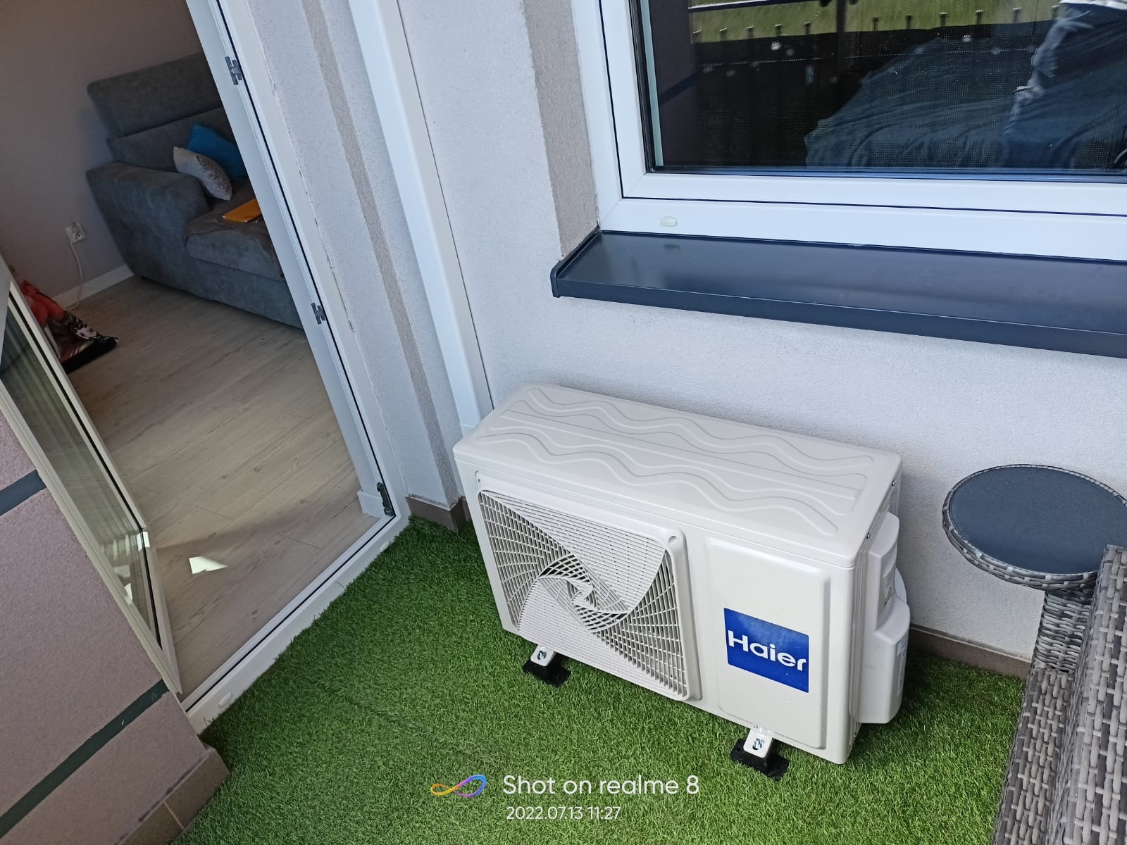 Zewnętrzna jednostka klimatyzatora Haier na balkonie z sztuczną trawą, widok z góry, widoczne okno i częściowo meble balkonowe oraz wnętrze mieszkania przez otwarte drzwi balkonowe.