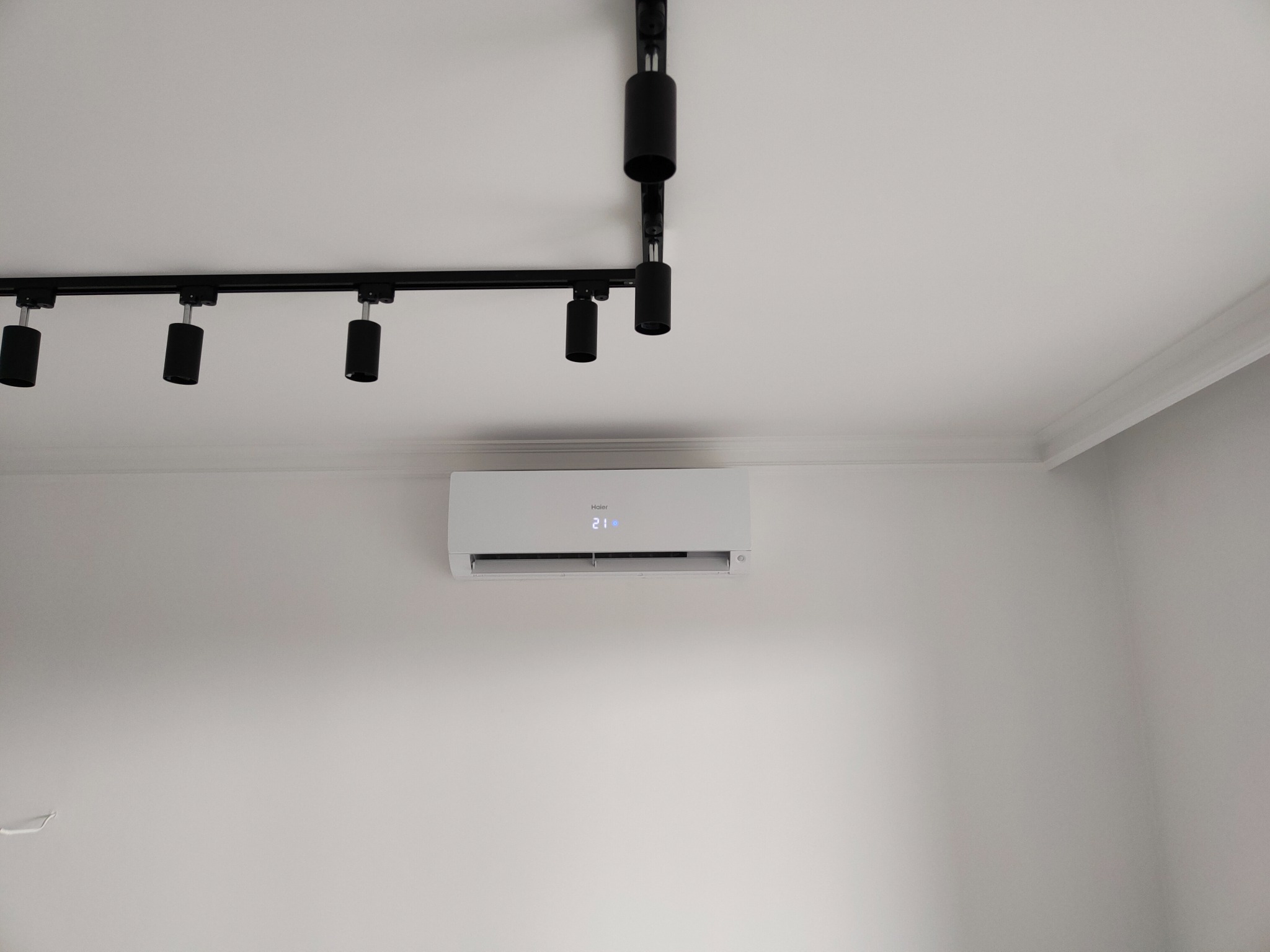 Zamontowany, biały klimatyzator Haier z wyświetlaczem temperatury 21 stopni Celsjusza na białej ścianie w pomieszczeniu w Katowicach, oświetlony czarnymi reflektorami sufitowymi.