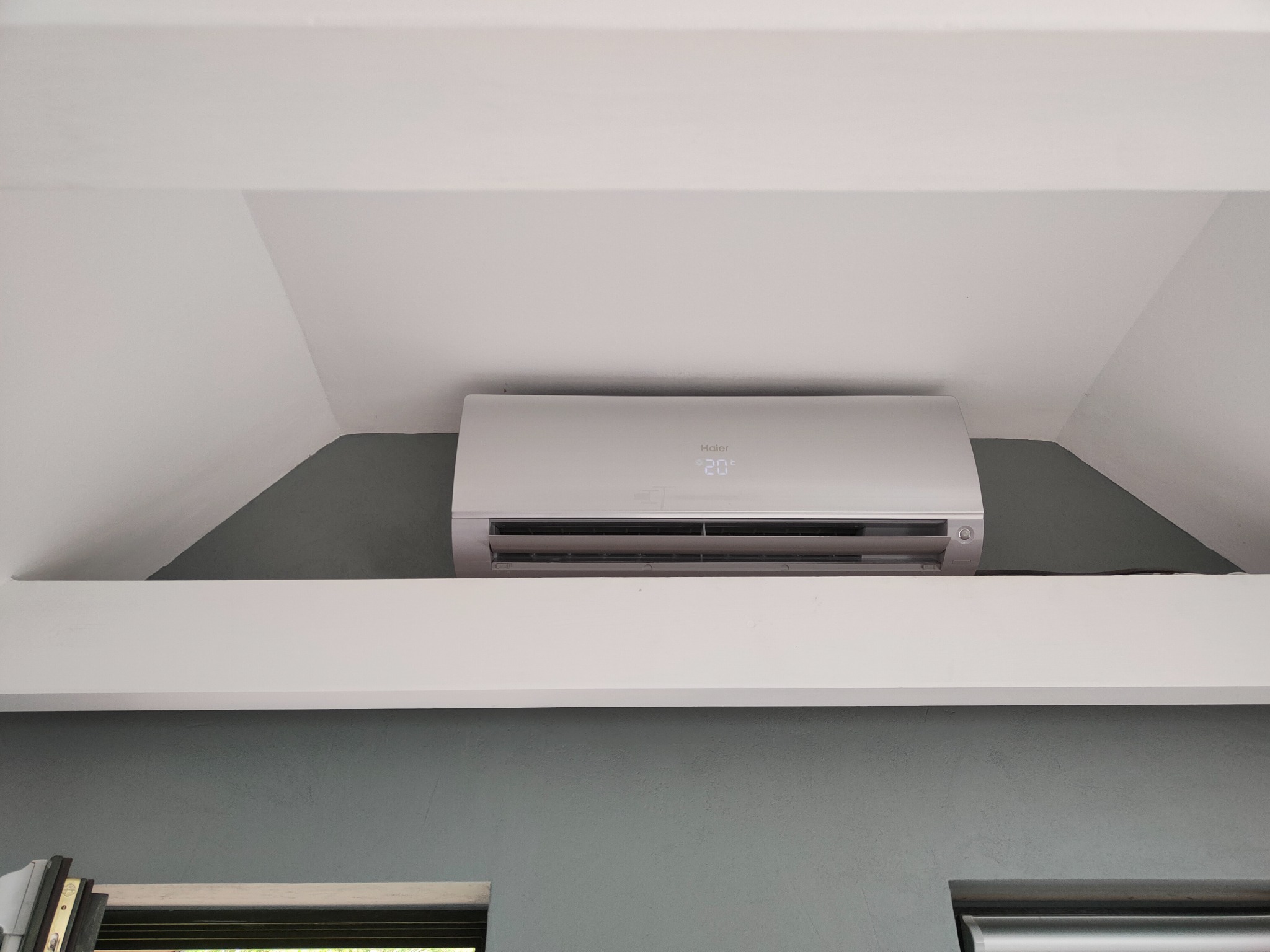 Biały klimatyzator Haier zamontowany w rogu pokoju na tle szarej ściany, wyświetlający temperaturę 20 stopni Celsjusza.