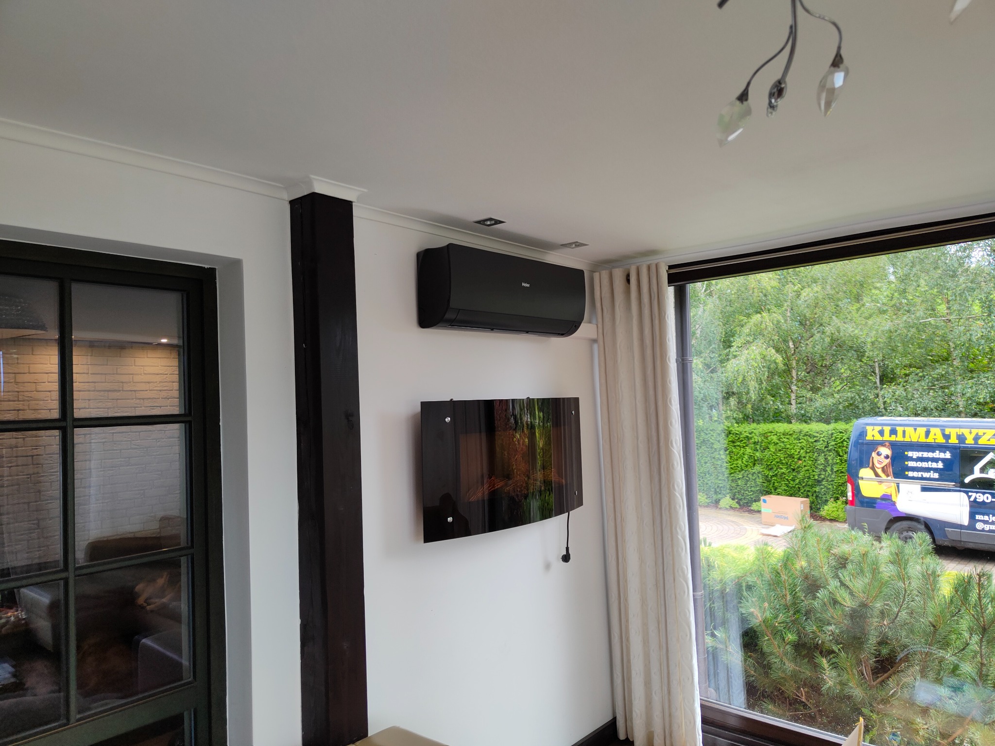 Czarny klimatyzator Haier zamontowany na białej ścianie nad czarnym, ozdobnym kominkiem elektrycznym w salonie z widokiem na podjazd, na którym stoi samochód serwisowy klimatyzacji.