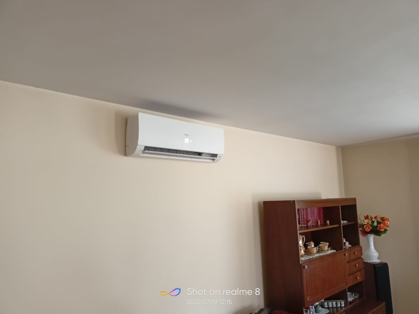 Biały klimatyzator Haier zamontowany na kremowej ścianie w pokoju mieszkalnym, widoczny brązowy kredens z dekoracjami.