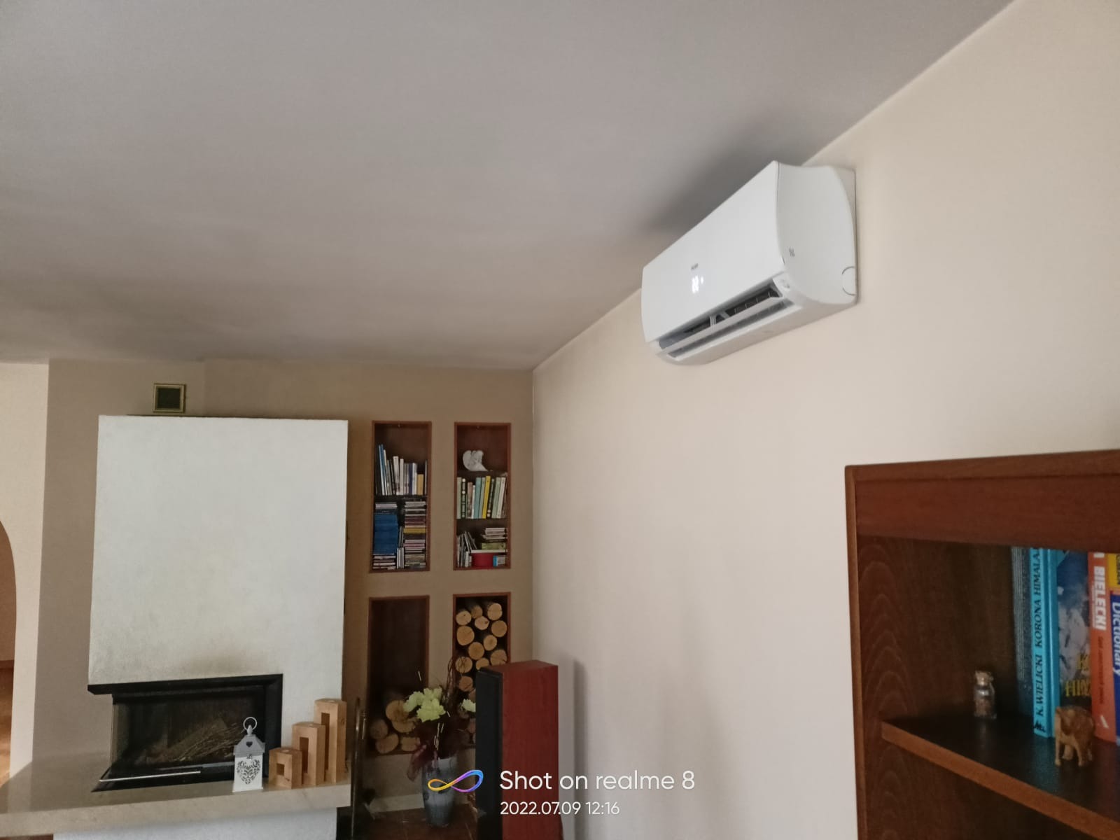 Biały klimatyzator ścienny w salonie z kominkiem i regałami na książki, widok z dołu.