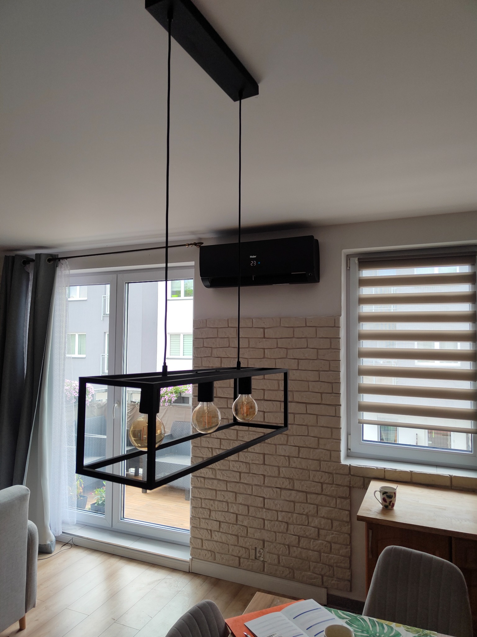 Nowoczesny czarny klimatyzator ścienny Haier zamontowany w salonie nad oknem balkonowym, widoczny designerski żyrandol z żarówkami Edisona.