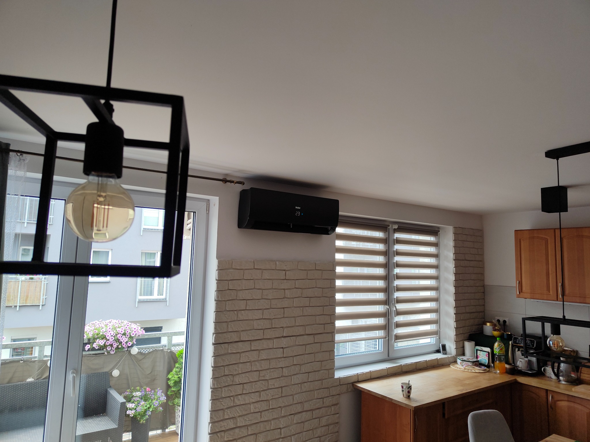 Czarny klimatyzator ścienny zamontowany nad oknem z żaluzjami w jasnej kuchni z ceglaną ścianą i nowoczesnym oświetleniem.