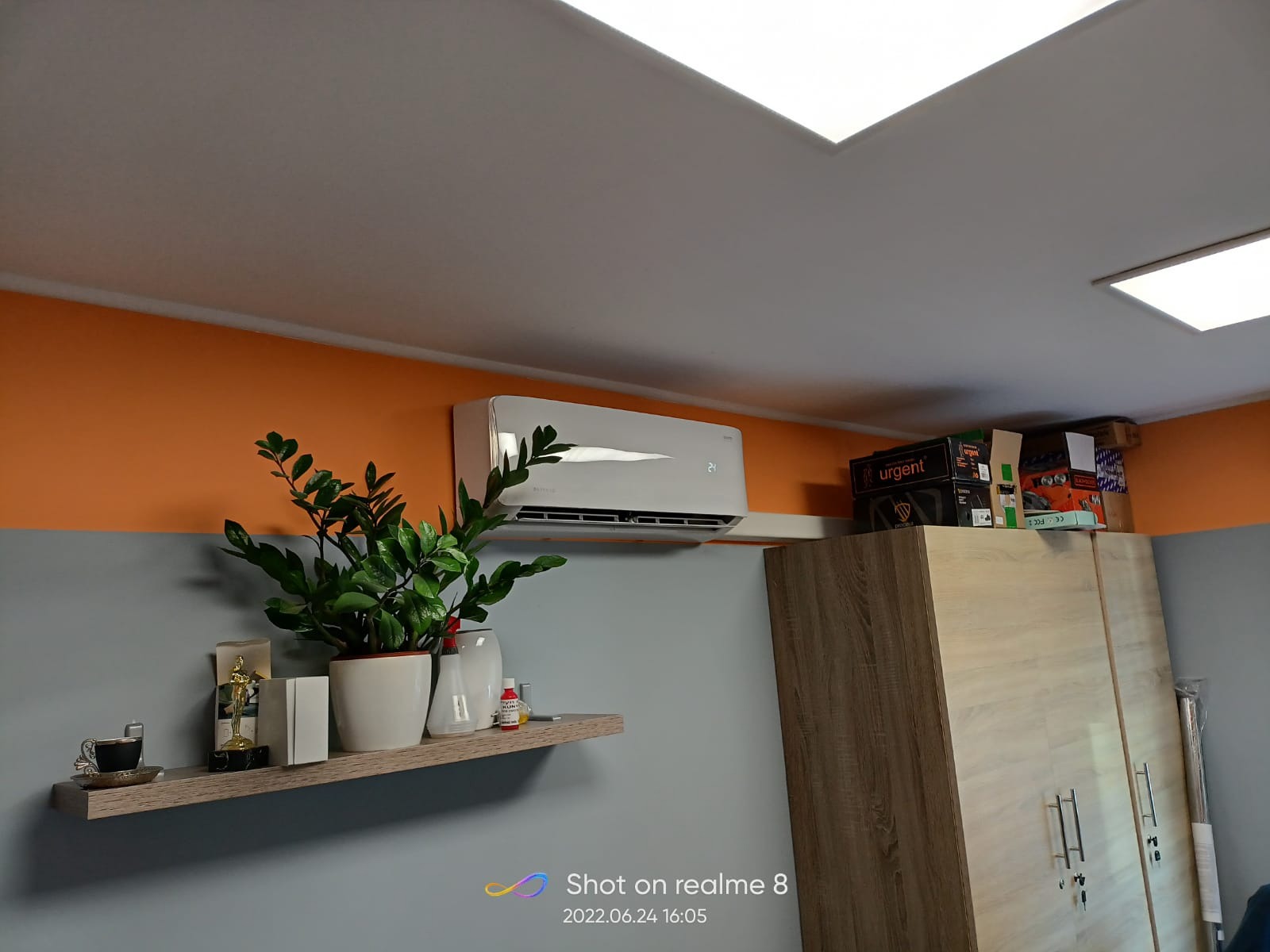 Biały klimatyzator ścienny zamontowany w pokoju z pomarańczowo-szarą ścianą, obok szafy i półki z rośliną doniczkową i dekoracjami.