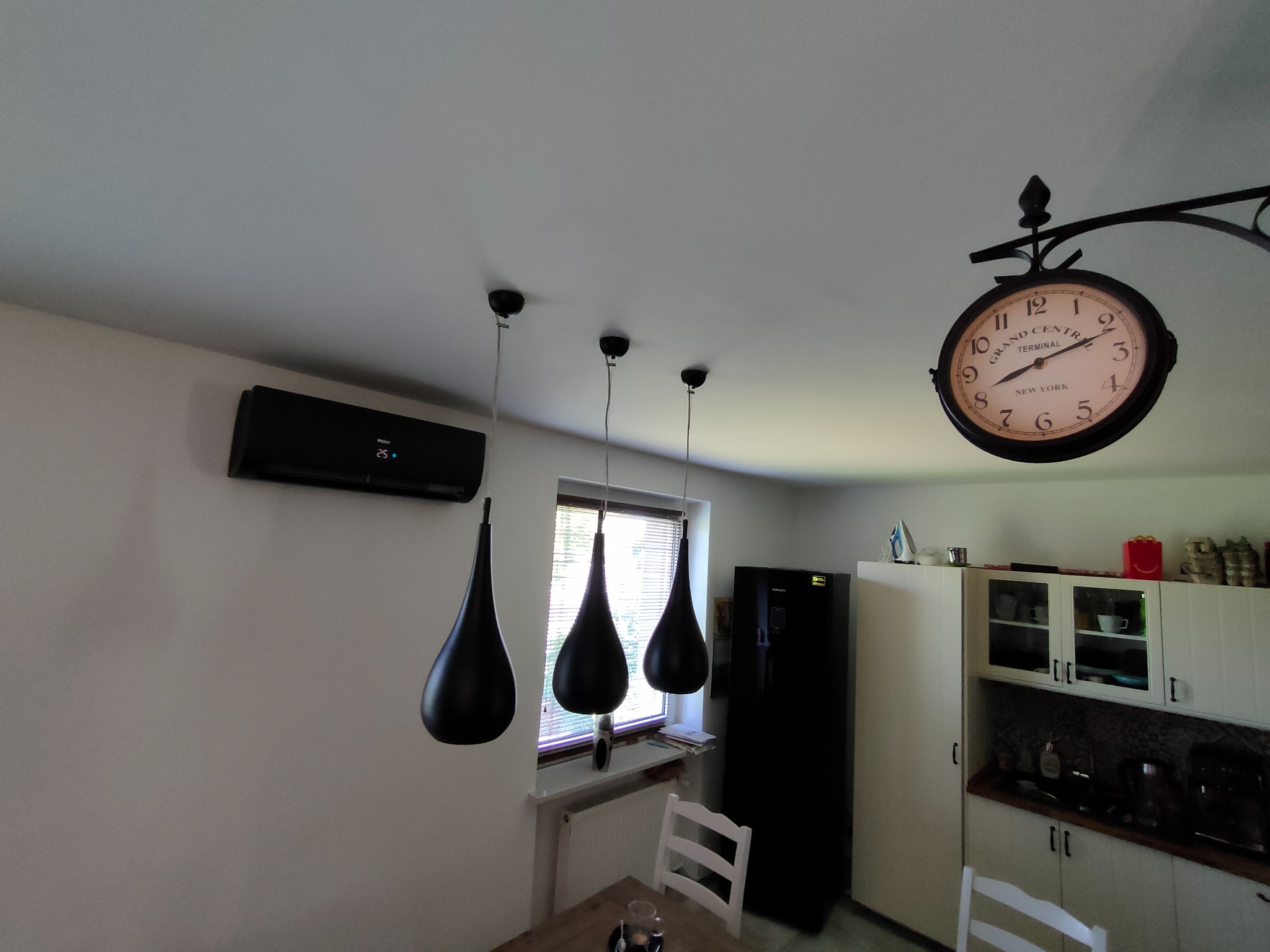 Wnętrze kuchni z zamontowanym czarnym klimatyzatorem ściennym marki Haier, trzema czarnymi lampami wiszącymi o kształcie łzy, wiszącym zegarem w stylu retro oraz czarną lodówką Samsung.
