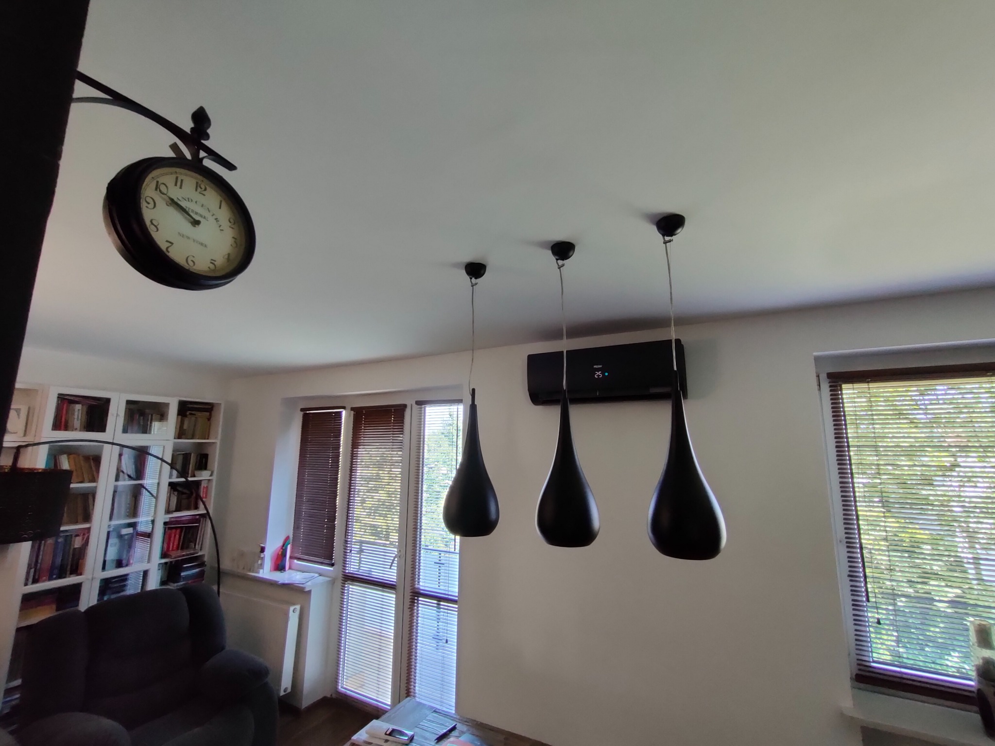 Czarny klimatyzator ścienny zamontowany nad trzema wiszącymi lampami w kształcie łezki, widok z dołu, na ścianie zegar i okna z żaluzjami.