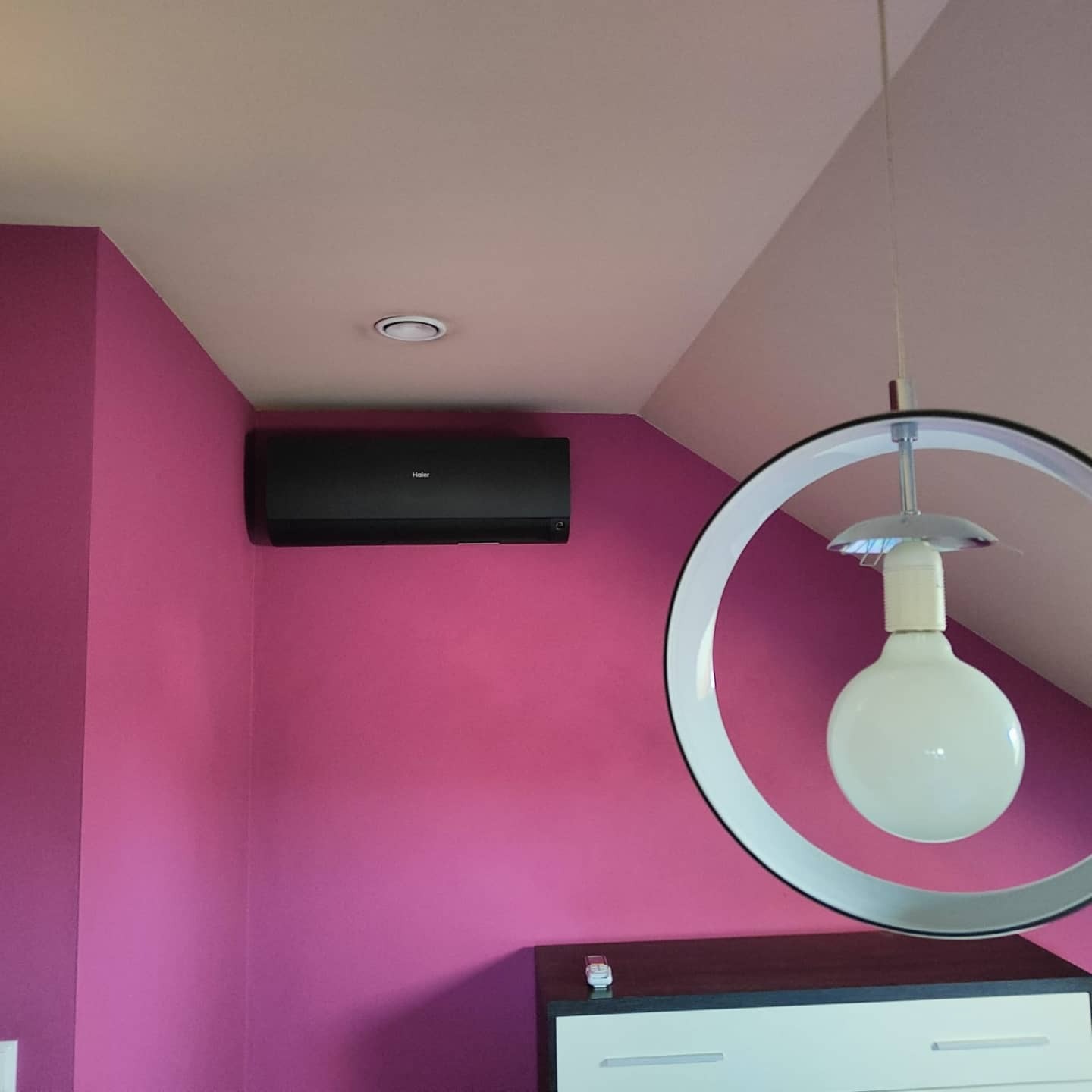 Czarny klimatyzator Haier na różowej ścianie w pokoju na poddaszu, widoczna lampa sufitowa z okrągłym kloszem i biała komoda.
