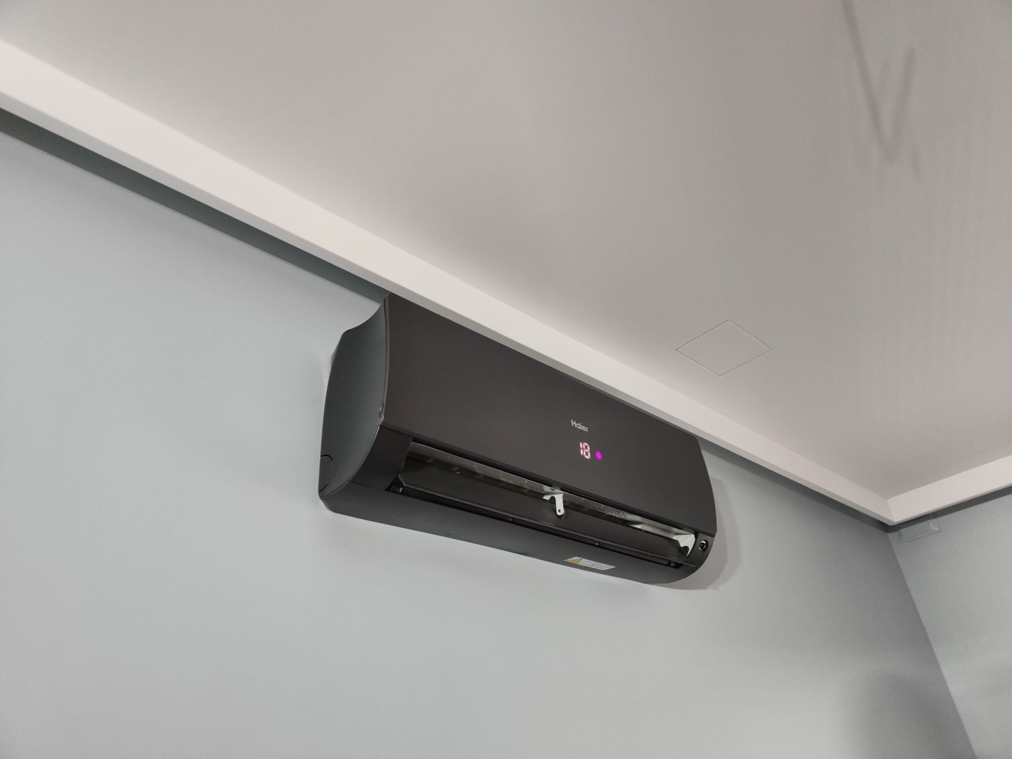 Czarny klimatyzator Haier zamontowany na szarej ścianie w pomieszczeniu z białym sufitem i listwą przysufitową. Widoczny wyświetlacz z temperaturą 18 stopni.
