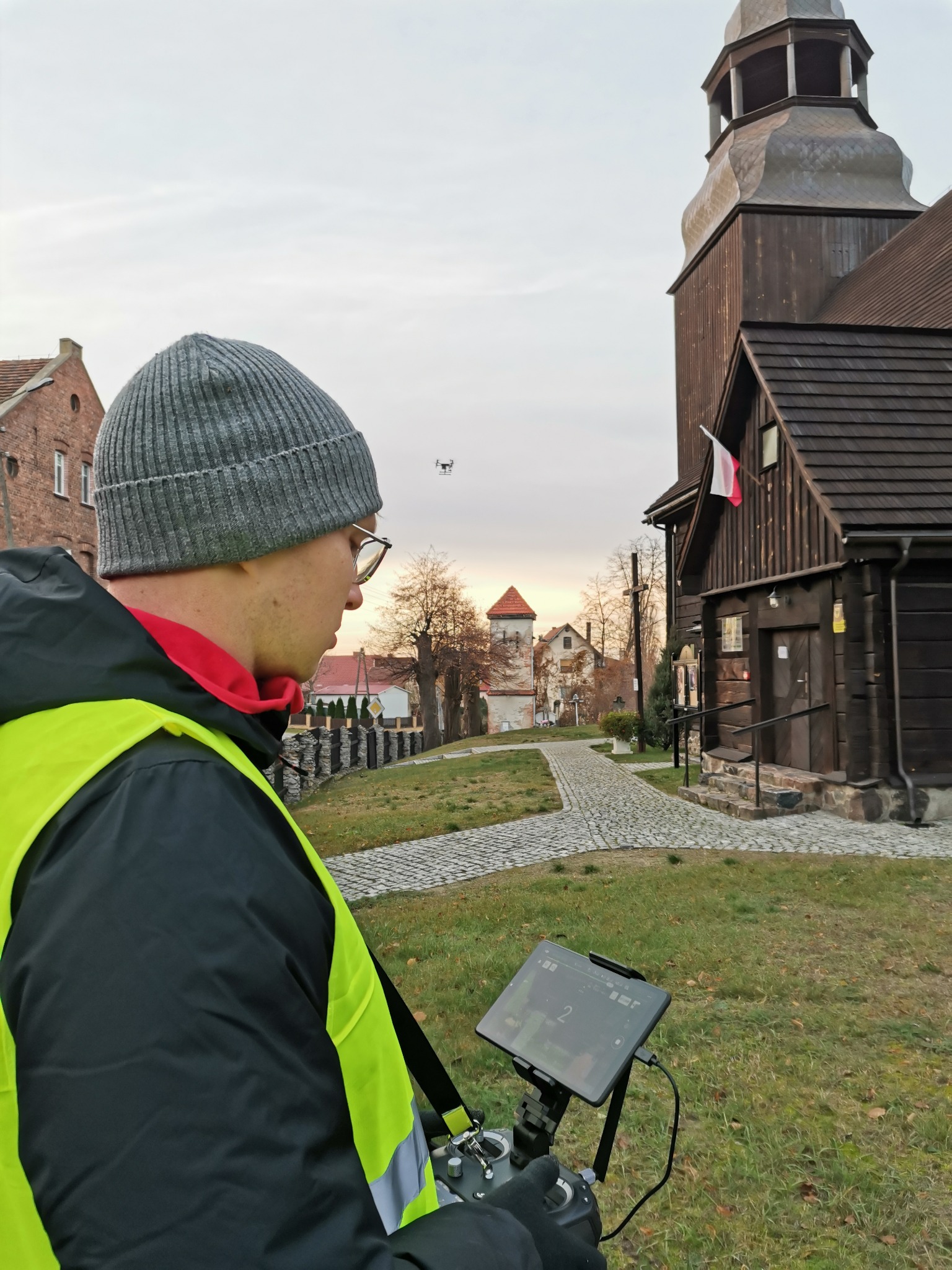 Mężczyzna w kamizelce odblaskowej i czapce, obsługujący drona w pobliżu drewnianego kościoła z polską flagą, prawdopodobnie wykonujący pomiary.