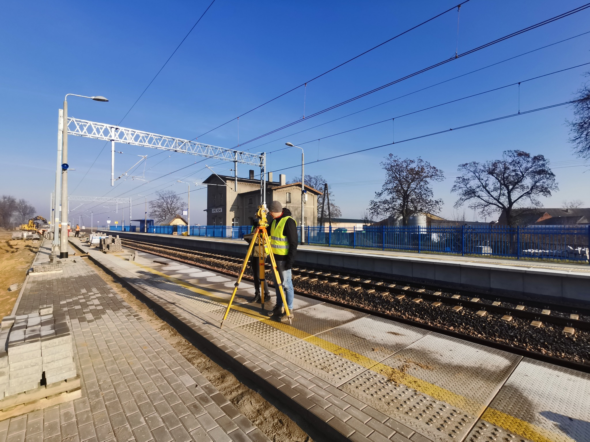 Praca geodezyjna na stacji kolejowej w Zawoni: pomiary wykonywane przy użyciu niwelatora na tle torów i budynku stacyjnego.