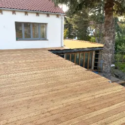 Drewniany taras w trakcie budowy, widoczne deski, drabina i fragment domu z ogrodem w tle.