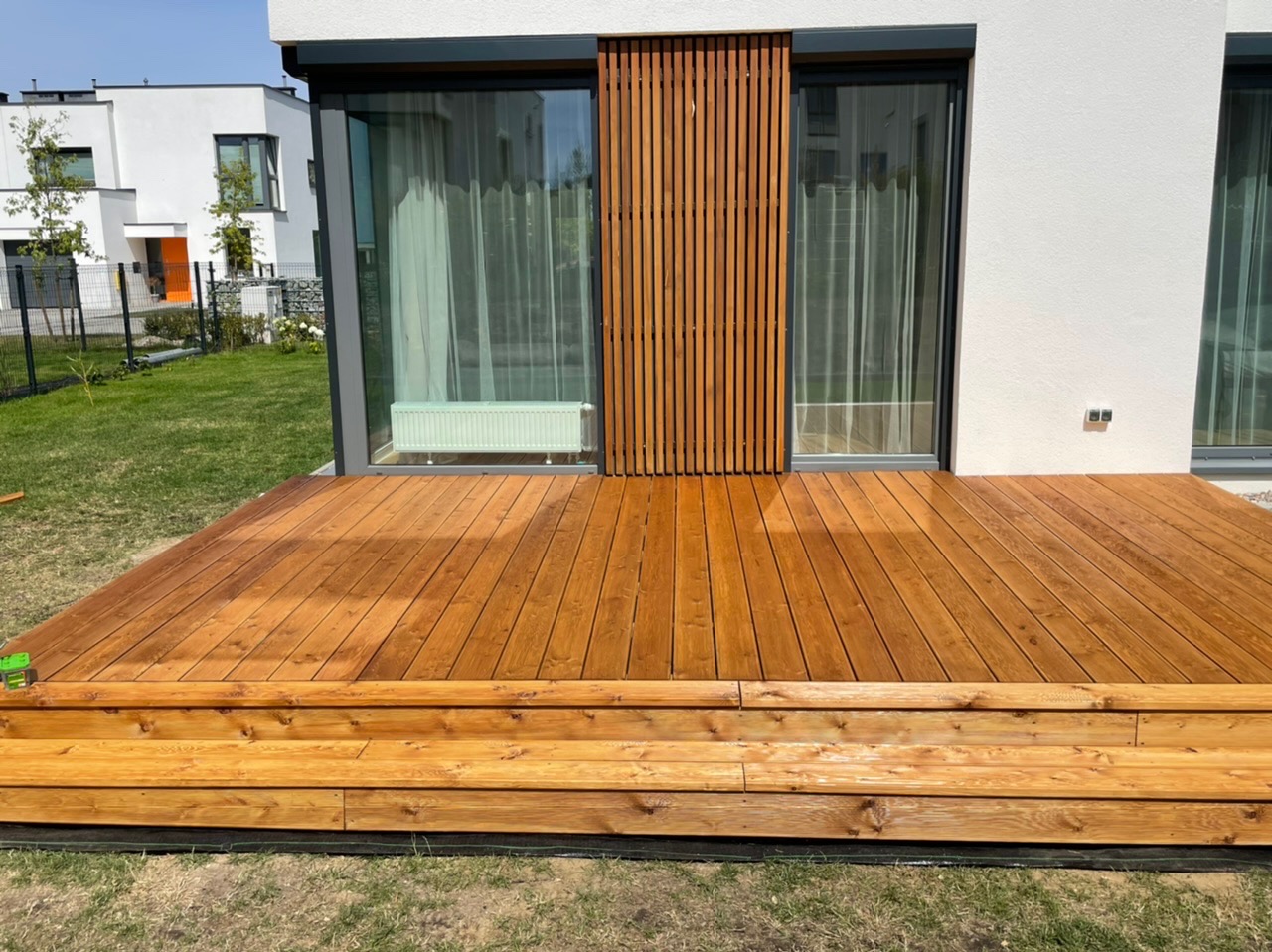 Nowoczesny taras z drewna o ciepłym odcieniu brązu, przylegający do białego domu z dużymi oknami i pionowymi drewnianymi panelami, widok na trawnik.