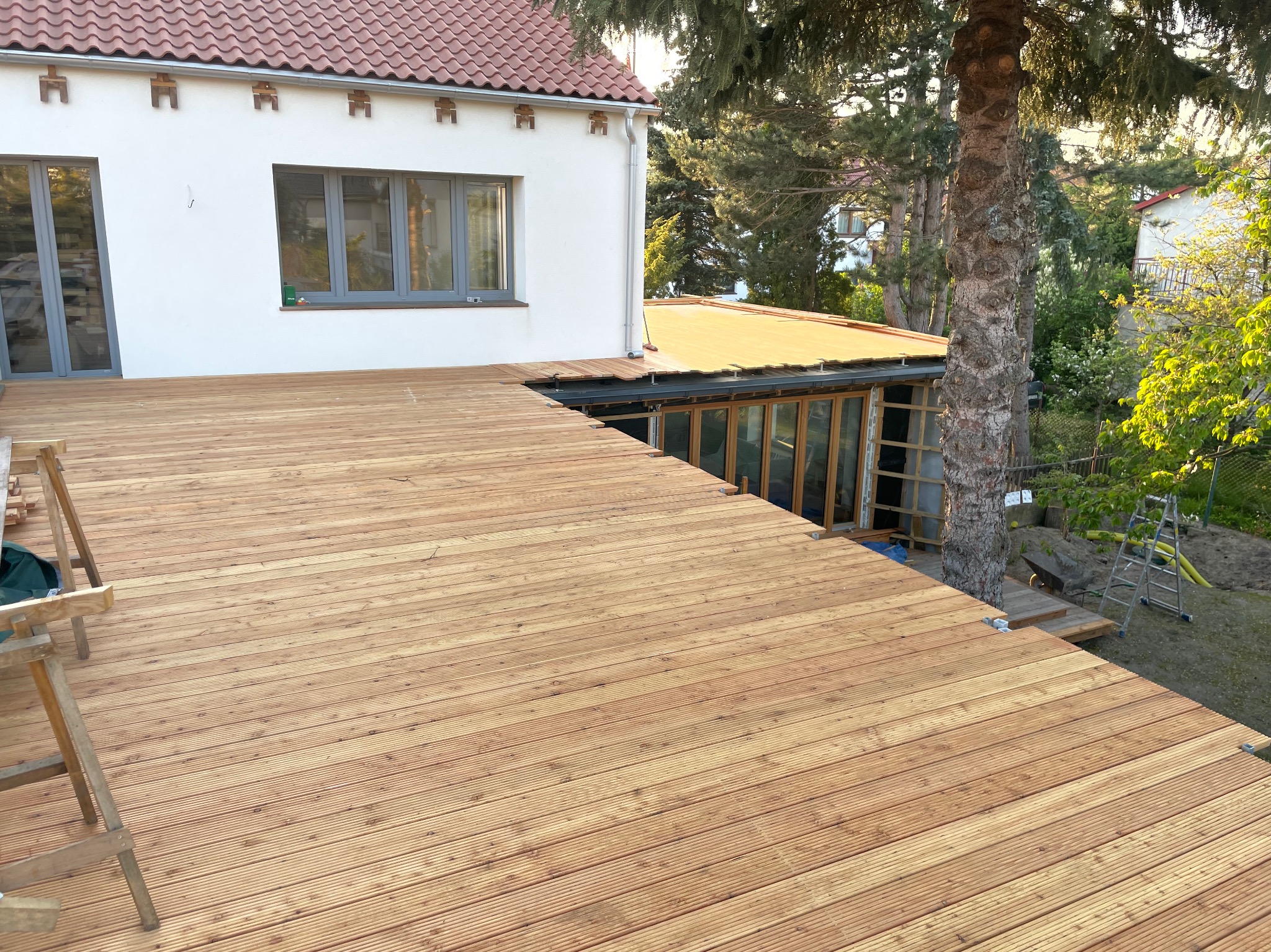 Drewniany taras w trakcie budowy, widoczne deski, drabina i fragment domu z ogrodem w tle.