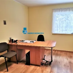 Wnętrze biura z drewnianym biurkiem, krzesłem, komputerem stacjonarnym, oknem z firanką i logo agencji nieruchomości na ścianie.