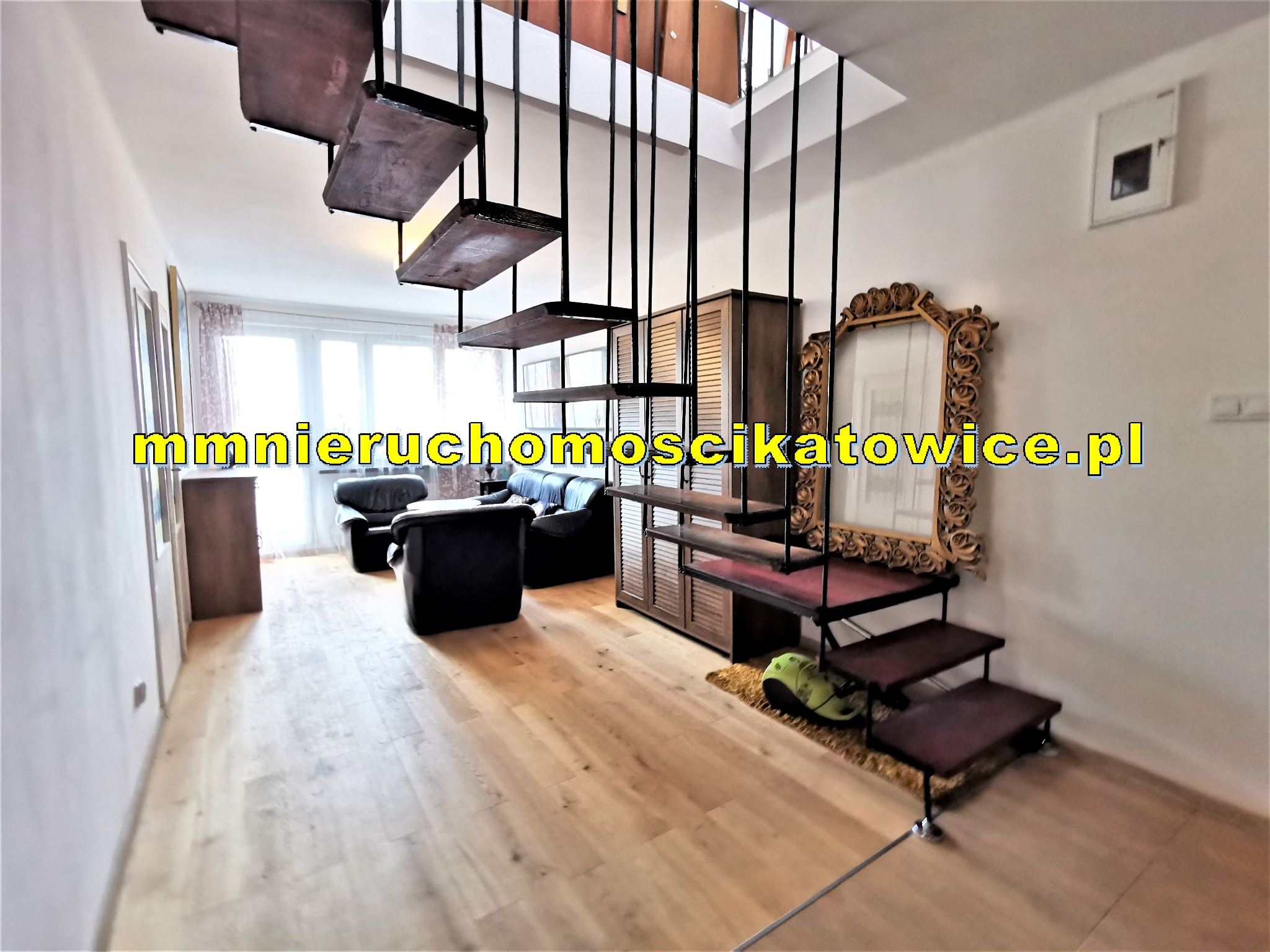 Jasne wnętrze mieszkania z drewnianą podłogą, czarnymi schodami na metalowych prętach, ozdobnym lustrem w złotej ramie i czarnymi skórzanymi sofami w tle.