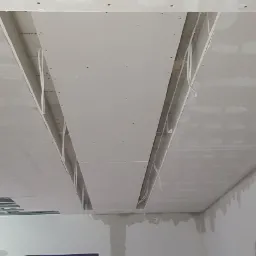 Montaż sufitu podwieszanego z płyt kartonowo-gipsowych, widoczne kable elektryczne w przestrzeni między sufitem a płytami, częściowo wykończone łączenia płyt.