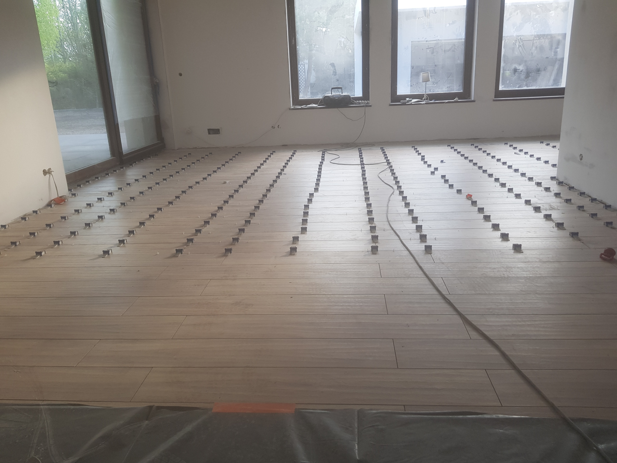 Układanie płytek podłogowych w dużym pokoju z widocznymi systemami poziomowania, jasne płytki imitujące drewno, przygotowane do fugowania.