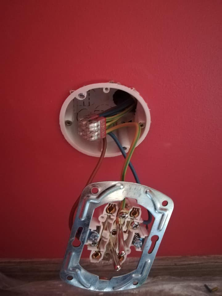 Otwarta puszka elektryczna w czerwonej ścianie z widocznymi przewodami i metalową ramką gniazdka, etap prac instalacyjnych.