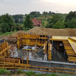 SZYC-BUD - Wykop pod fundamenty domu jednorodzinnego z widocznym szalunkiem, zbrojeniem i betonem wylanym w części wykopu. W tle widoczny dom z czerwoną dachówką oraz zieleń.