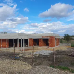 Surowy stan domu z czerwonej cegły z betonowym stropem w trakcie budowy, widok zewnętrzny na tle błękitnego nieba z chmurami. Ziemia wokół budynku pokryta gruzem i kamieniami, widoczne drewniane...