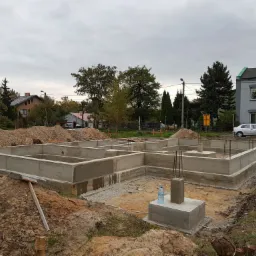 Surowe betonowe fundamenty domu jednorodzinnego z wystającymi prętami zbrojeniowymi, widoczne pryzmy ziemi i butelka wody na fundamencie, w tle zabudowa mieszkalna.