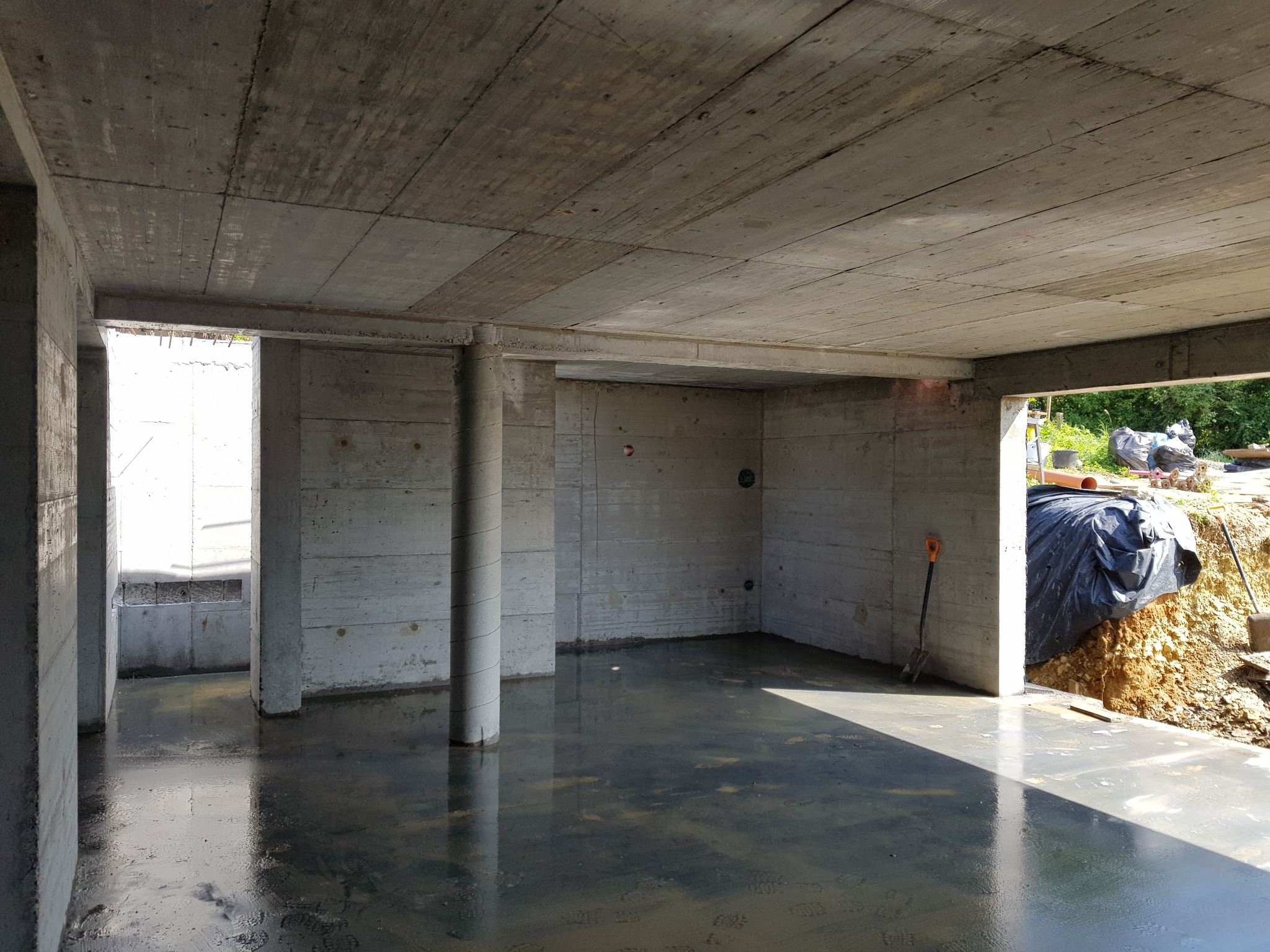 Surowe, żelbetowe wnętrze w trakcie budowy, zalaną posadzką i odciśniętym wzorem szalunku na ścianach i suficie, widoczna szpadel oparta o ścianę.