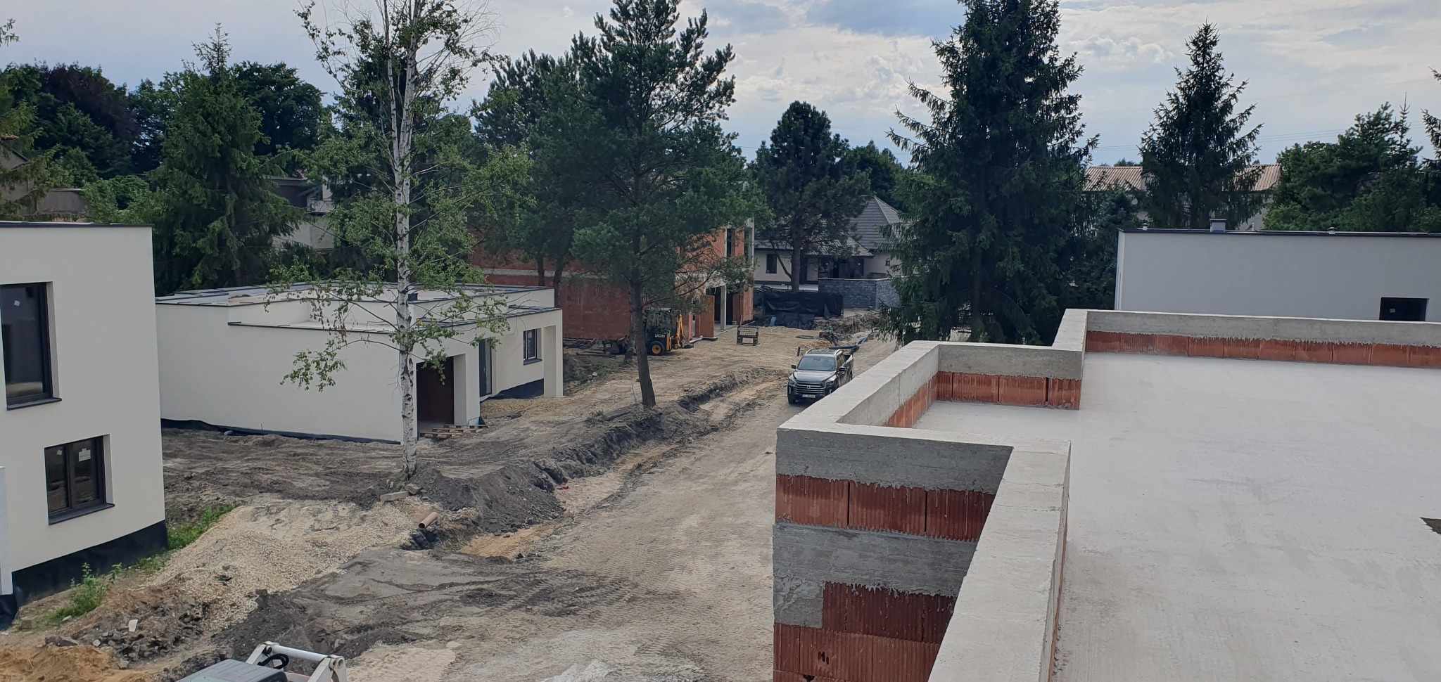 Wysoka perspektywa na plac budowy z surowym stanem budynków. Widoczne białe budynki z oknami, ceglane ściany w budowie i samochód na tle drzew.