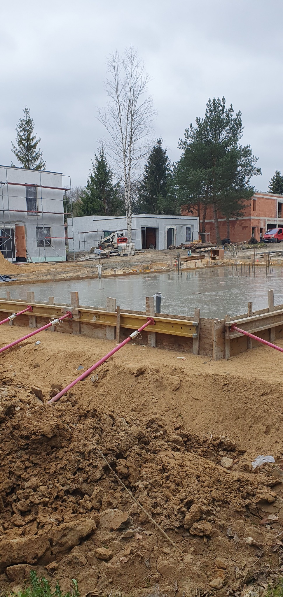 Wylany fundament z szalunkiem i podporami w Sosnowcu. W tle budynki w trakcie budowy i drzewa. Pochmurny dzień. Widoczne ślady prac ziemnych.