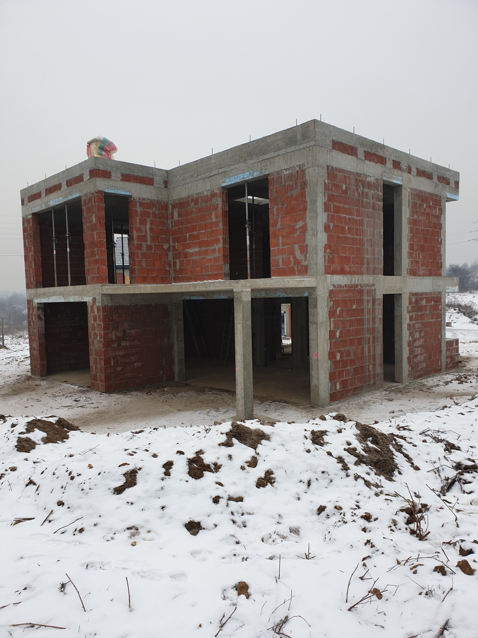Dwukondygnacyjny dom w stanie surowym otwartym, z czerwonej cegły i betonowych słupów, częściowo ośnieżony teren budowy, worek z zaprawą na dachu.
