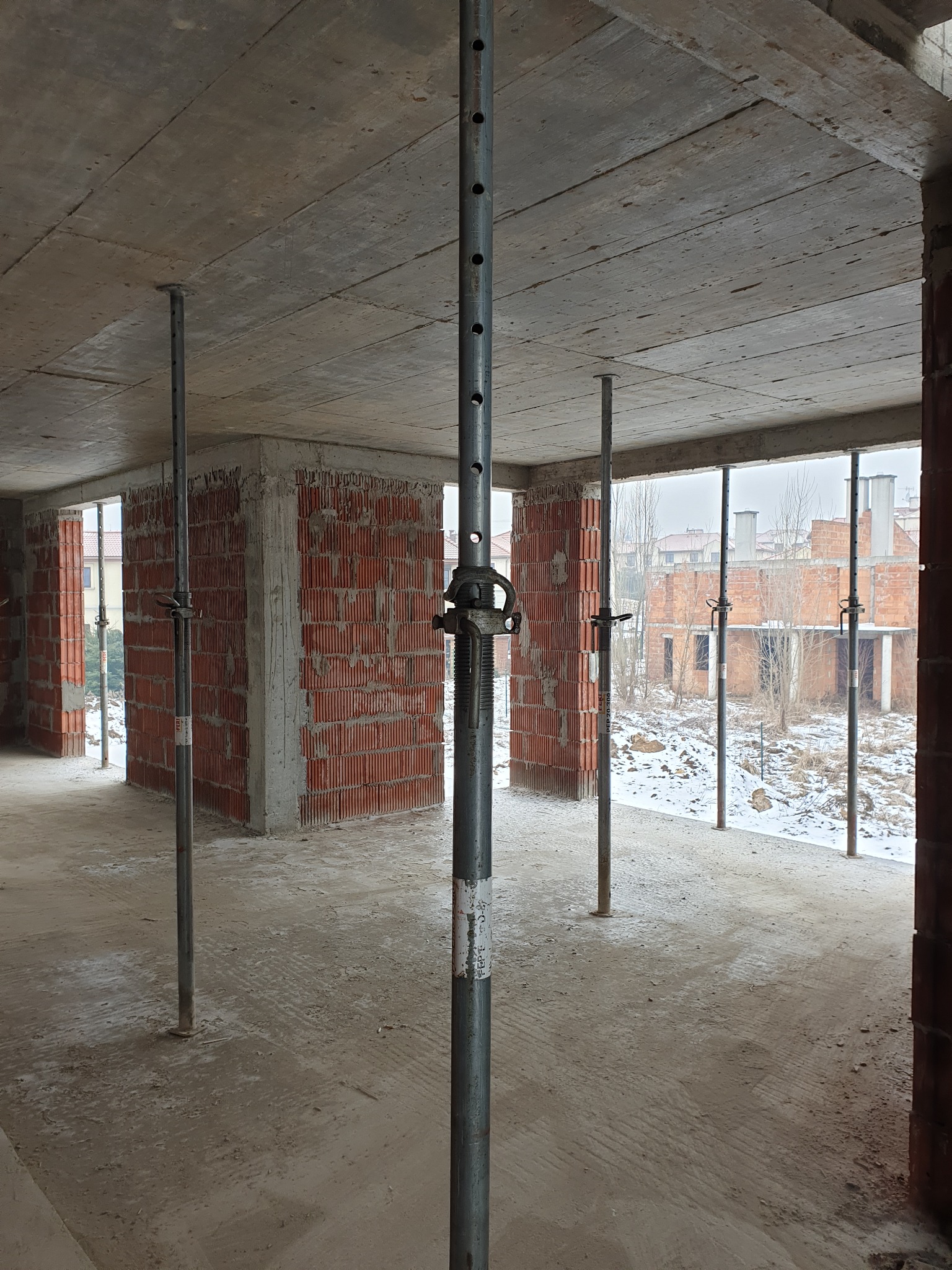 Wnętrze surowego budynku w trakcie budowy, widoczne ściany z czerwonej cegły, betonowy strop podparty stalowymi stemplami, za oknami zimowy krajobraz z innymi budynkami w budowie.