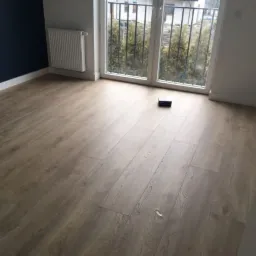 Jasne panele podłogowe ułożone w nowym mieszkaniu z balkonem, widoczne gniazdka elektryczne na ciemnej ścianie, grzejnik i czarny przedmiot na podłodze.