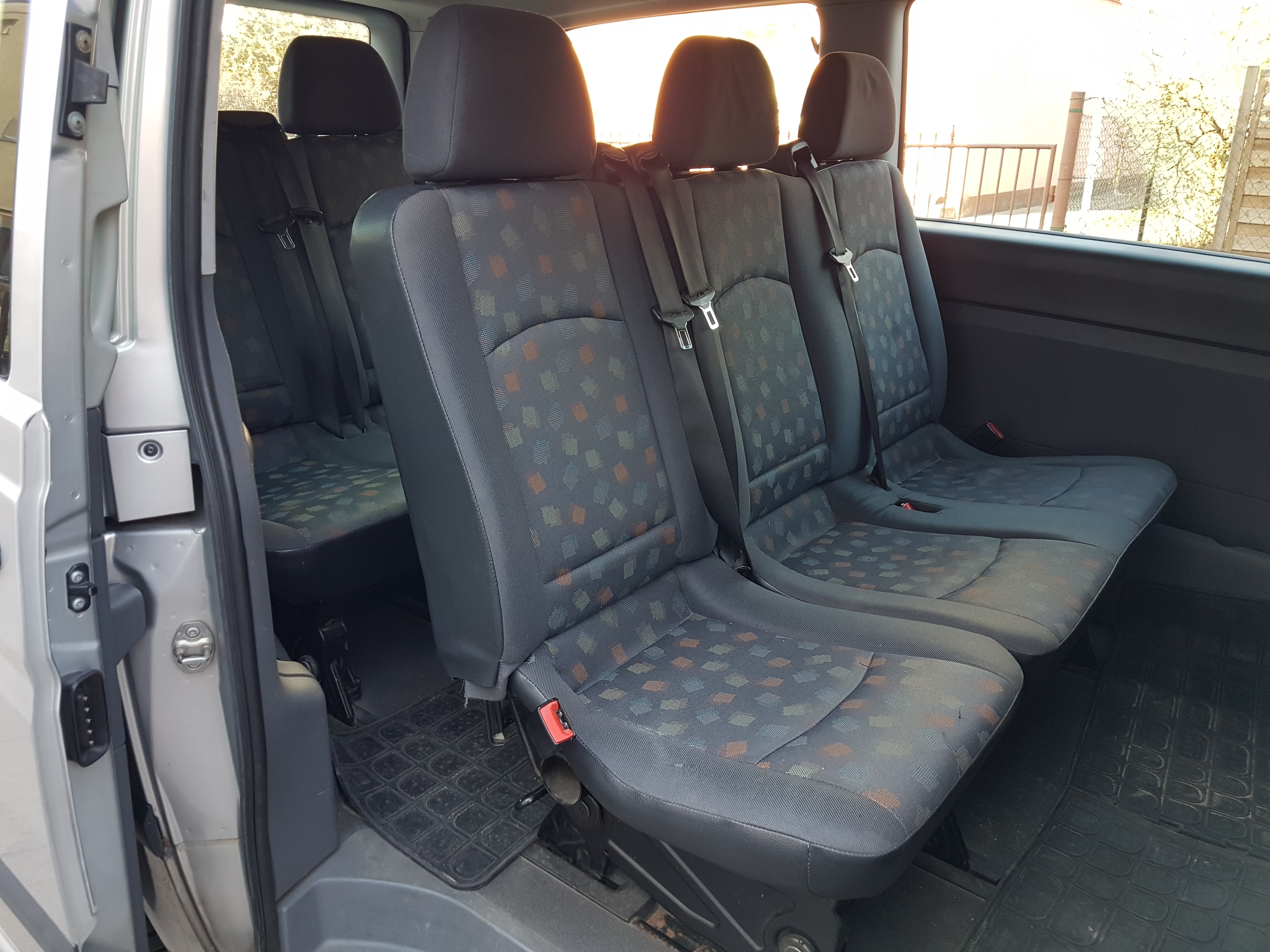 Wnętrze srebrnego busa z trzema rzędami foteli z ciemną tapicerką w kolorowe romby, widok od strony otwartych drzwi.