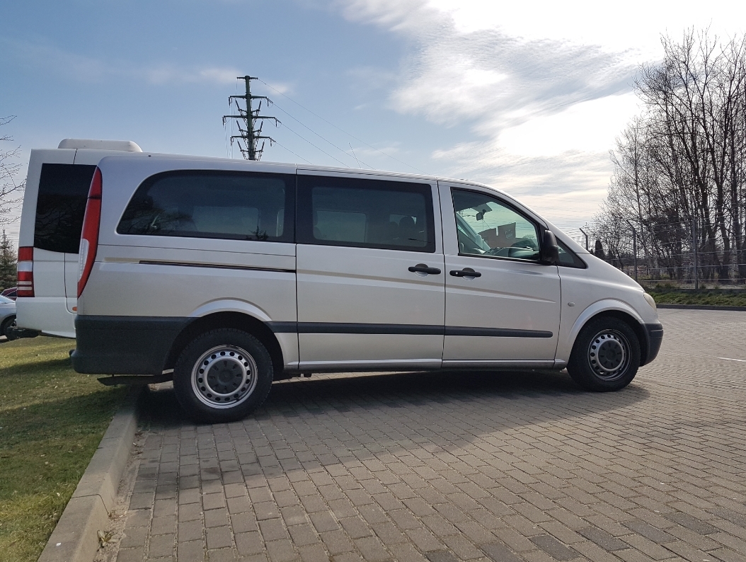 Jasnoszary van Mercedes Vito zaparkowany na kostce brukowej, widok z boku, zarys drugiego busa w tle, słupy wysokiego napięcia w oddali.