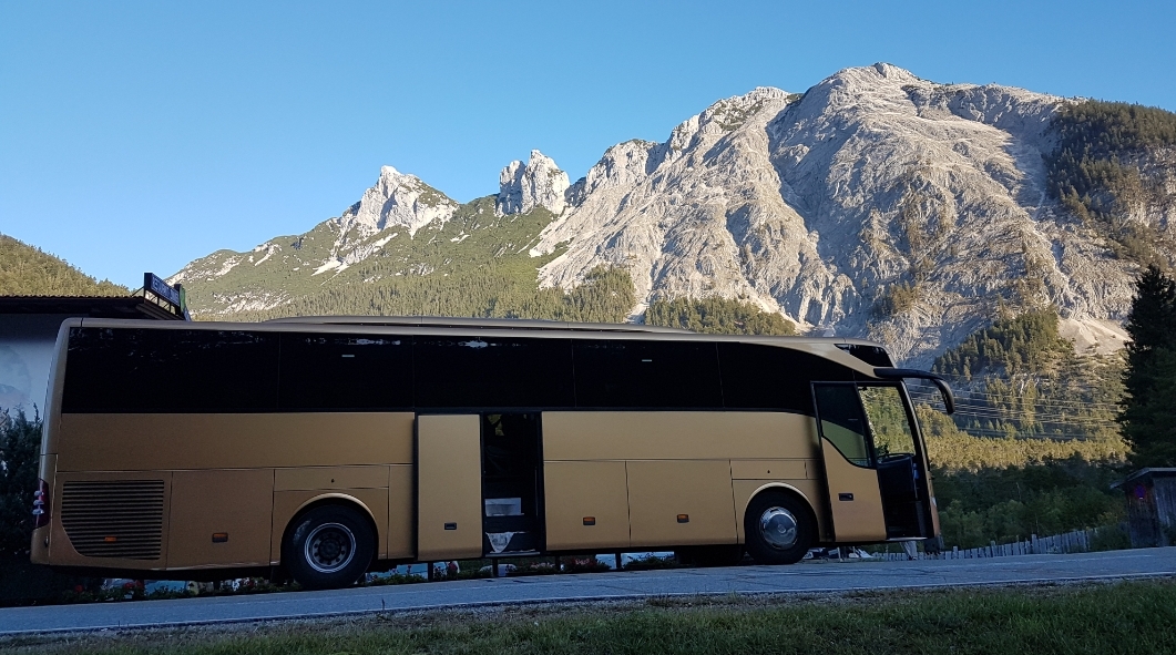 Złoty autokar turystyczny z otwartymi drzwiami na tle malowniczego górskiego krajobrazu z wyraźnymi szczytami i zielenią drzew.